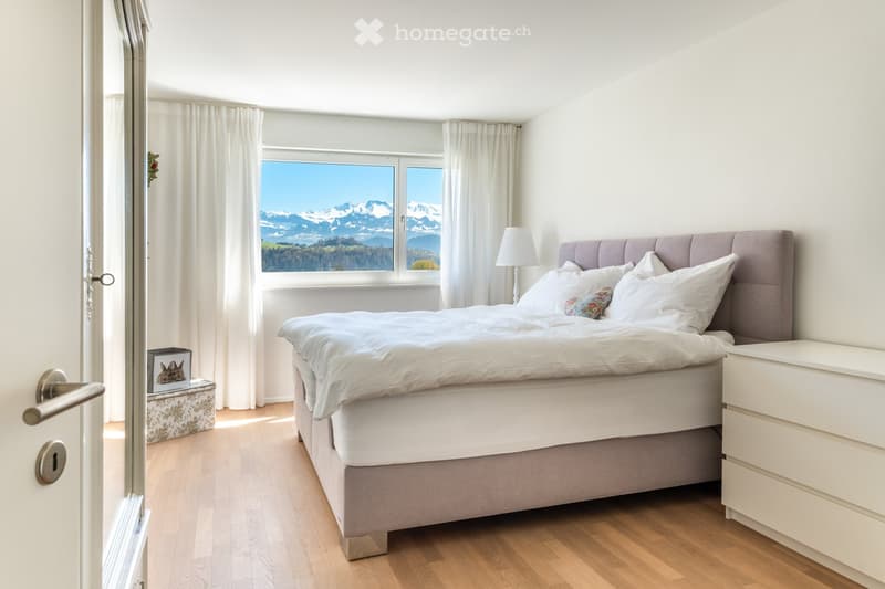 Bedroom. Bright bedroom with a large window offering a scenic mountain view, featuring a neatly made bed, a nightstand with a lamp, and a decorative picture frame on a small bench.
