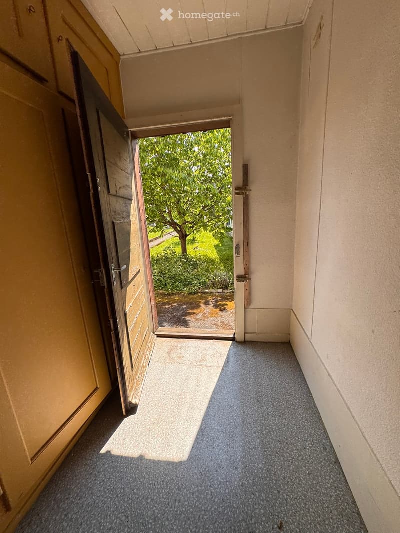 Corridor / Hallway. A narrow hallway with a grey carpeted floor, beige walls, and an open door leading to a garden area with trees and greenery.