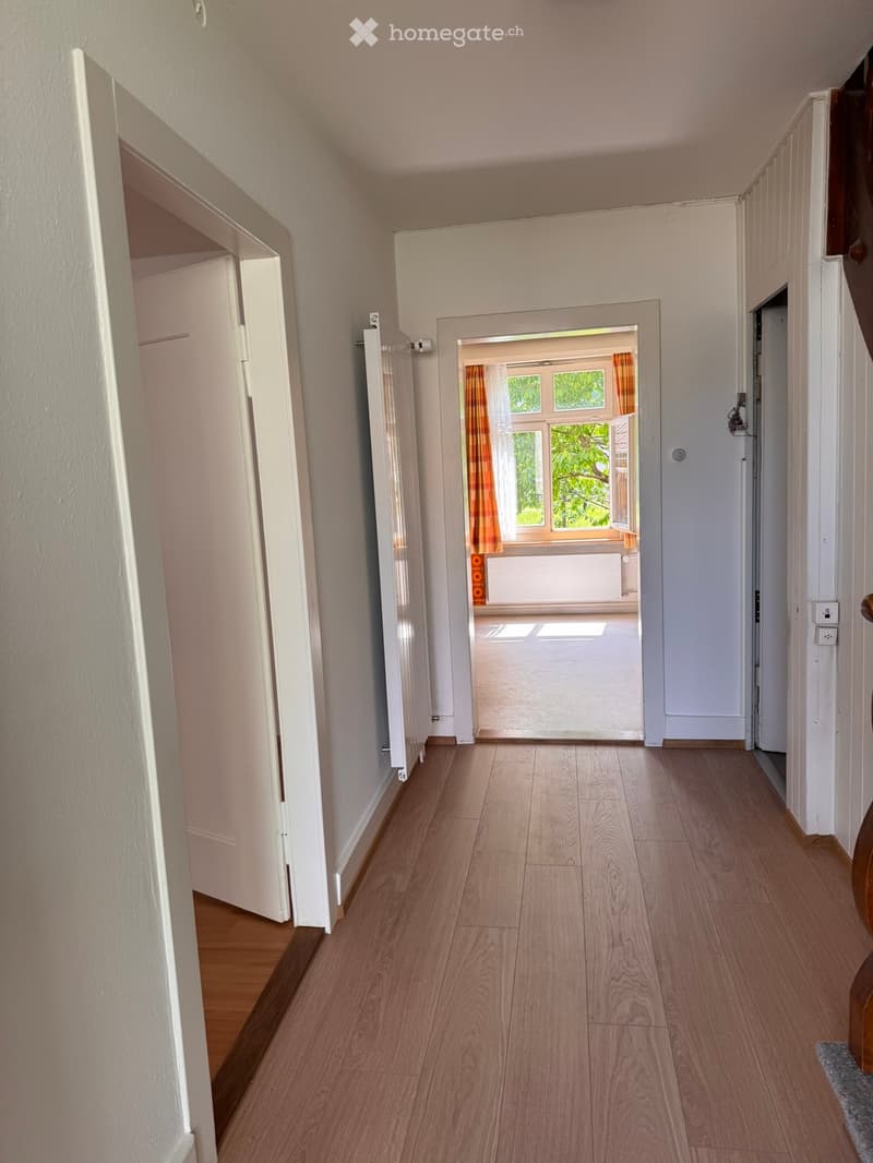 Corridor / Hallway. A clean, well-lit hallway with wooden flooring, white walls, and multiple doors leading to different rooms. Natural light streams in from a window at the end of the hallway.