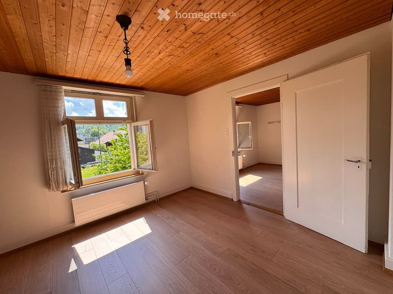 Unfurnished Room. Empty room with wooden flooring, white walls, large window with curtains, and a visible adjacent room with a radiator and a door.