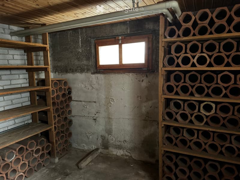 Unfurnished Room. Empty storage room with exposed brick walls, wooden shelving, and a small window. Shelves hold neatly stacked cylindrical objects, likely pipes or logs.