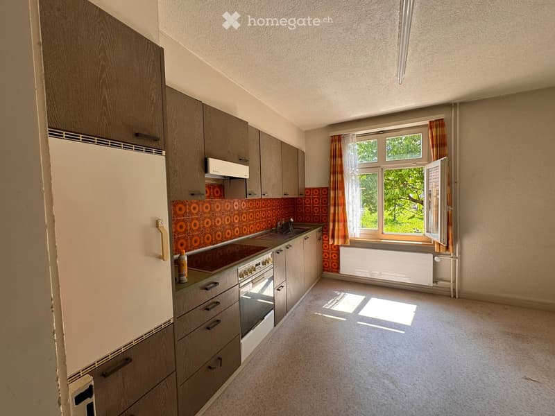 Kitchen. A small, unfurnished kitchen with wooden cabinets, a white refrigerator, a sink, and an oven. The space features a retro orange backsplash and a window with striped curtains.