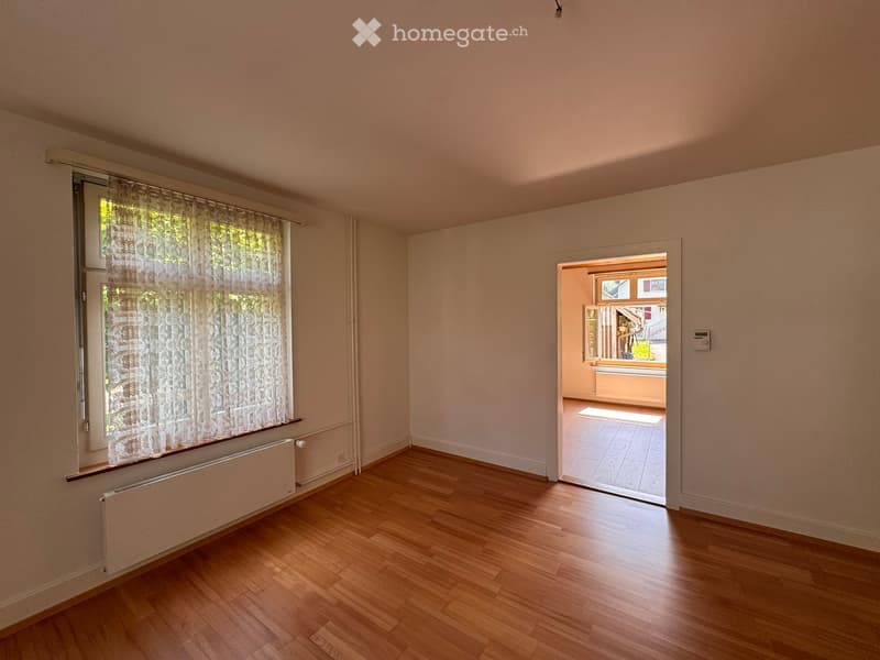 Unfurnished Room. Empty room with wooden flooring, light-colored walls, and a large window with white curtains. A radiator is visible below the window, and an open doorway leads to another room with tiled flooring.