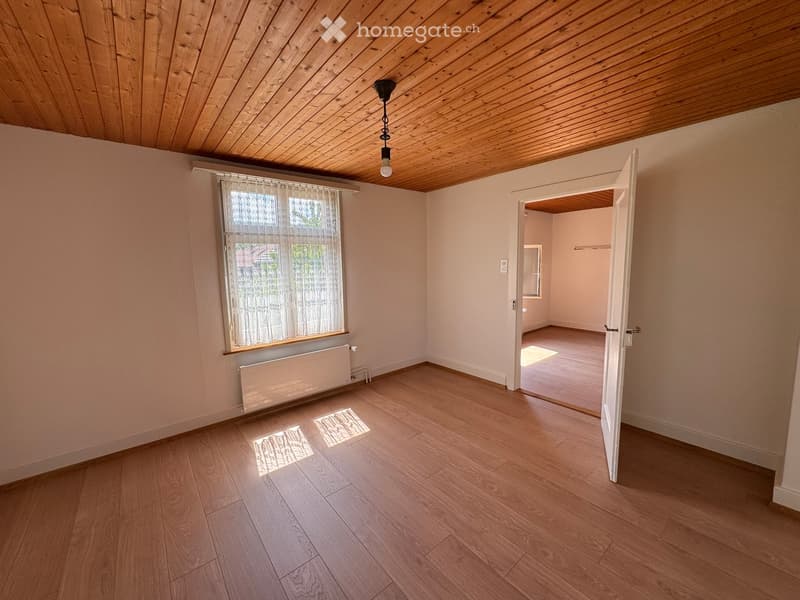 Unfurnished Room. Empty room with wooden flooring, light-colored walls, and a window with white curtains. A radiator is visible under the window, and an open door leads to another room.