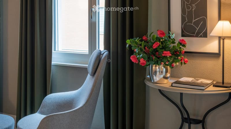 Furnished Room. A modern, cozy room with a grey upholstered chair, a small round table with a vase of pink flowers, books, and a lamp on a side table. The room features large windows with dark curtains and a framed abstract artwork on the wall.