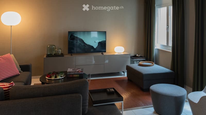 Living Room. Modern living room with a grey sofa, coffee table, ottoman, and a television on a sleek cabinet. The room features warm lighting from floor and table lamps, decorative jars, and books on the coffee table.