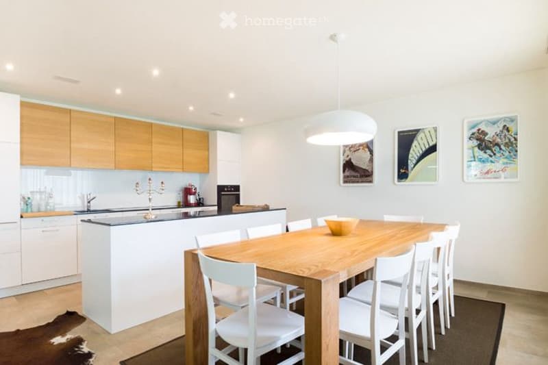 Furnished Room. Open-plan kitchen and dining area with modern white cabinetry, a central island, and a wooden dining table surrounded by white chairs. A hanging pendant light illuminates the space, and decorative posters adorn the walls.
