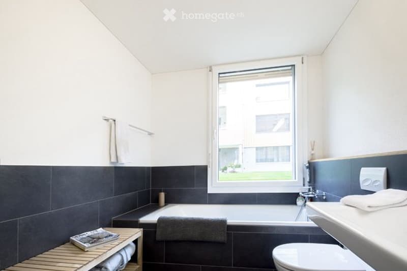 Bathroom. Modern bathroom with a bathtub, toilet, sink, and a wooden shelf holding towels and a magazine. The walls are tiled in dark gray with white upper sections, and there is a large window offering a view of the outside.