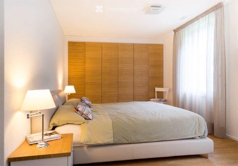 Bedroom. A modern bedroom with a large bed, nightstand, lamp, and a wooden wardrobe. The room features light-colored walls, wooden flooring, and a large window with curtains.