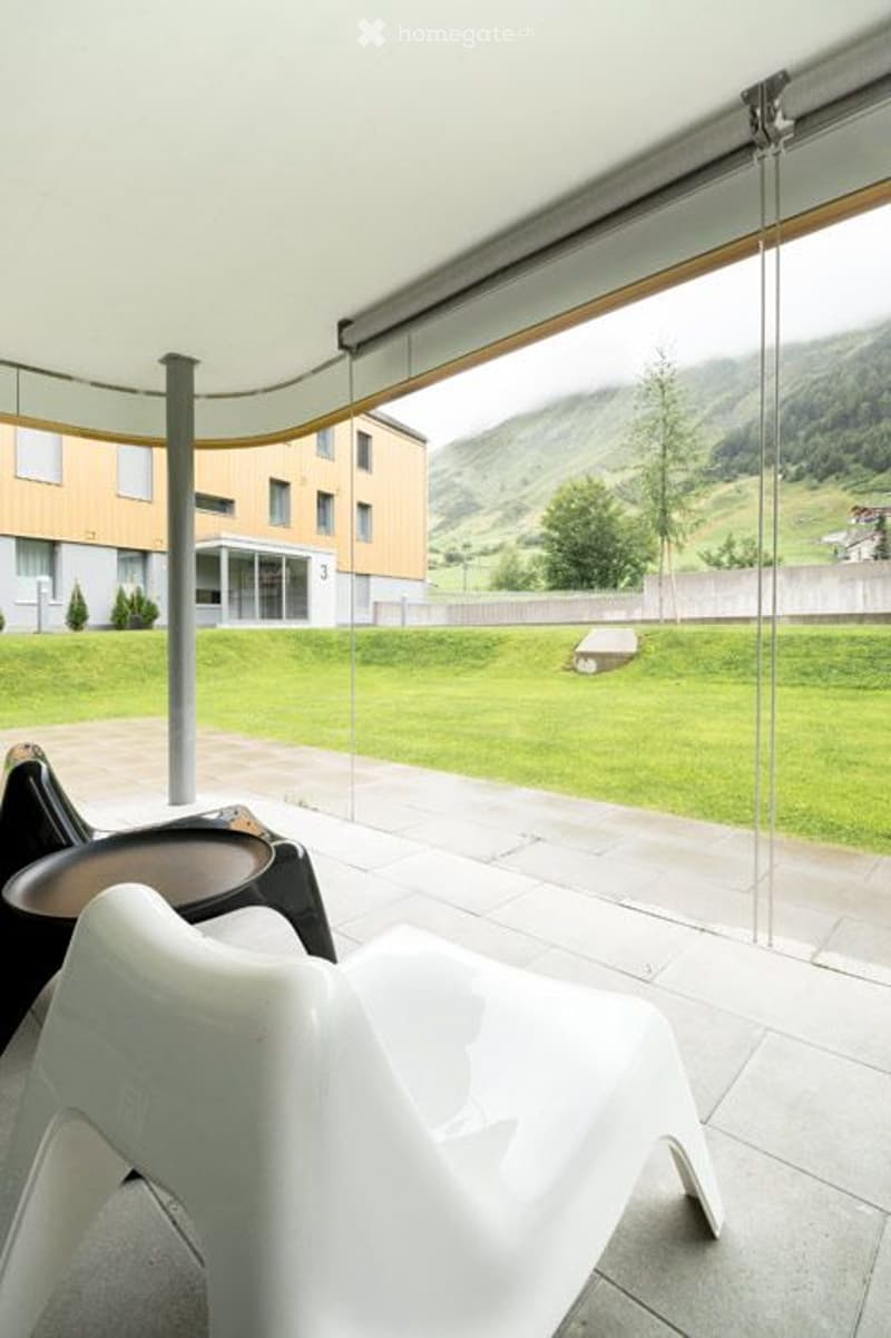 Balcony / Terrace. Modern balcony with large glass walls offering a scenic mountain view, featuring white plastic chairs and a small table.