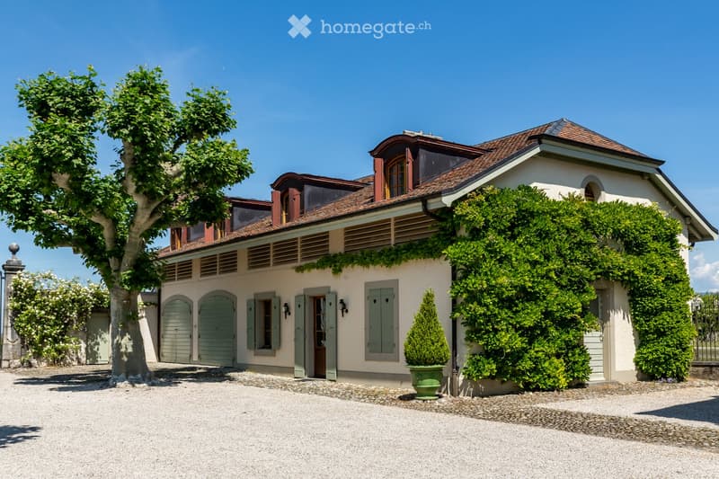 House. Two-story house with a red-tiled roof, white walls, green shutters, and a gated driveway. Features include a garage, a tree, and well-maintained landscaping.