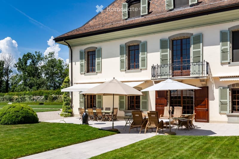 House. House, white walls, brown roof, green windows, balcony, garden, patio with dining area and umbrellas, hedges, trees