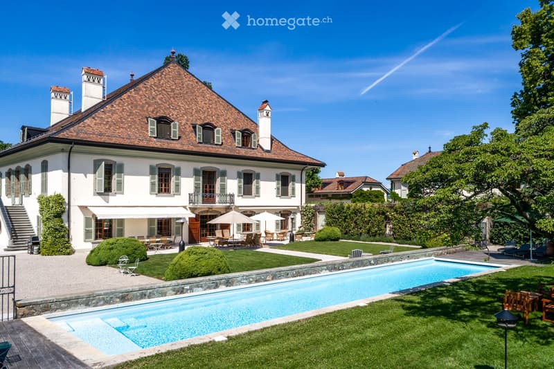House. A large, elegant two-story house with a red-tiled roof and white walls, surrounded by a well-maintained garden, a swimming pool, and outdoor seating areas with umbrellas and tables.