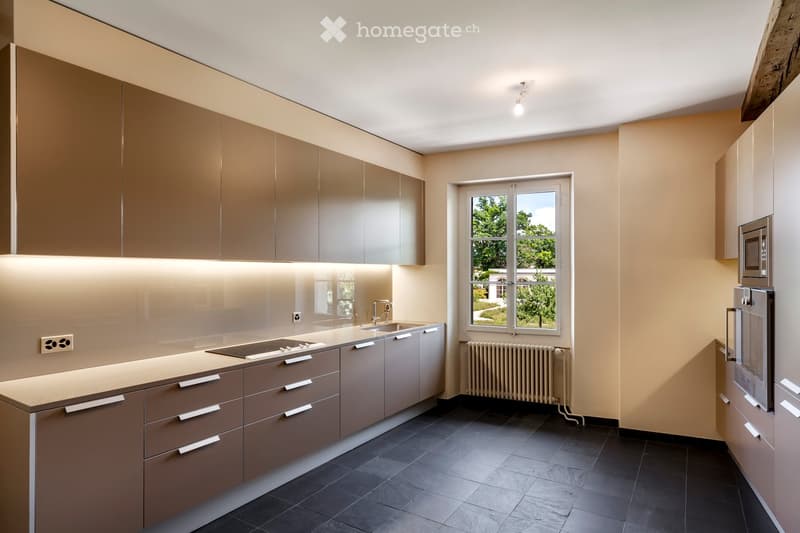 Kitchen. Modern kitchen with light brown cabinets, dark tiled floor, and a large window overlooking a garden. Features include a sink, microwave, and radiator.