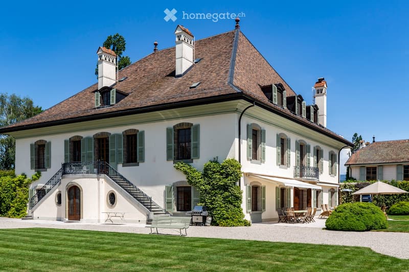 House. A large, elegant two-story house with a classic European design, featuring a brown tiled roof, white walls, green shutters, and multiple chimneys. The house has a well-maintained garden with a gravel driveway, a bench, and outdoor seating area.