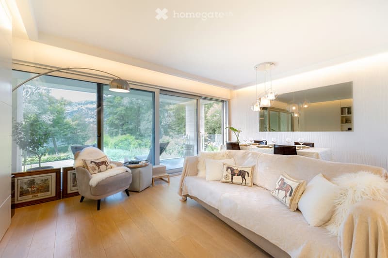 Living Room. Modern living room with large windows overlooking a green landscape, featuring a white sofa with decorative pillows, a matching armchair, a dining table with wine glasses, pendant lights, and framed artwork on the wall.