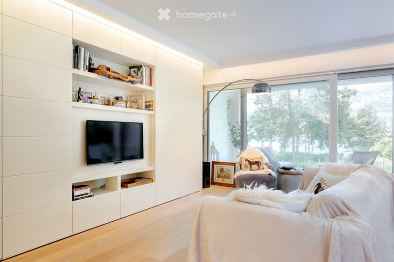 Living Room. Modern living room with white built-in shelving, a large flat-screen TV, a cream-colored sofa, an armchair, and a floor lamp. Large windows offer a view of greenery outside.