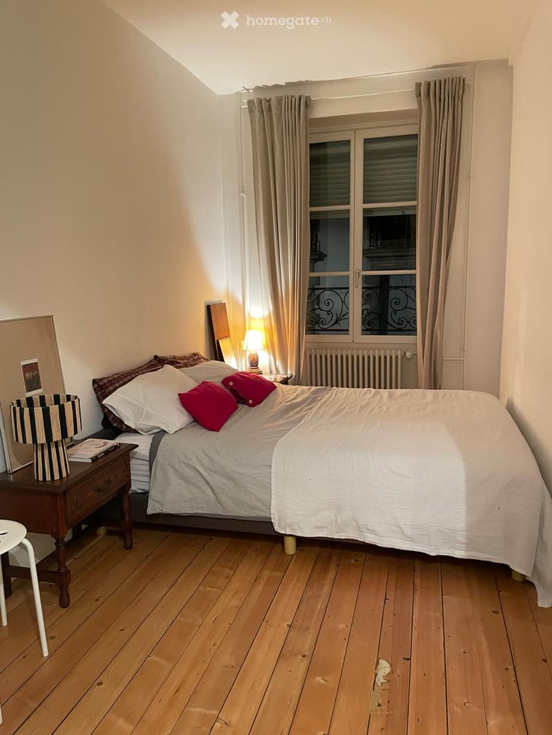 Bedroom. Cozy bedroom with wooden flooring, a neatly made bed with white linens and red pillows, a wooden nightstand with a lamp, a mirror, and a small stool near the window.