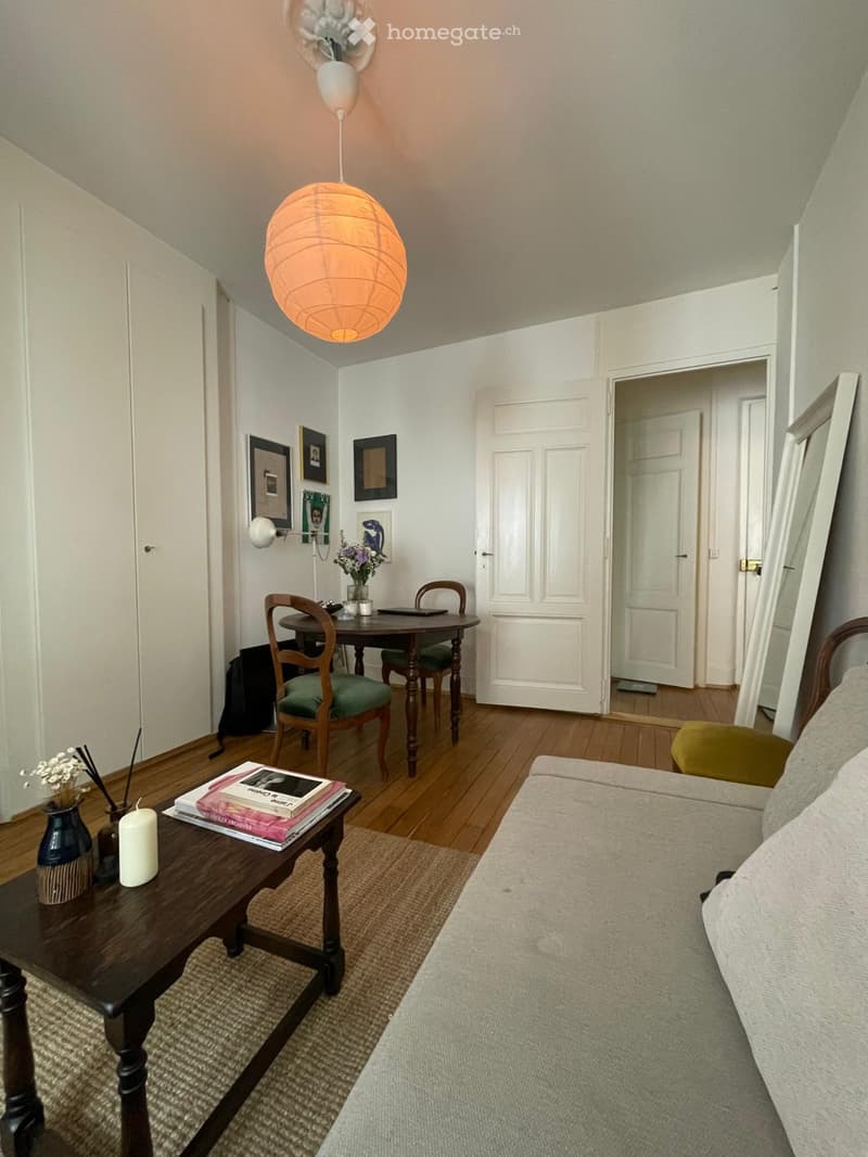 Furnished Room. A cozy living room with a sofa, coffee table, dining area with a table and chairs, and decorative items including books, a vase, and a candle. The room features wooden flooring and white walls.