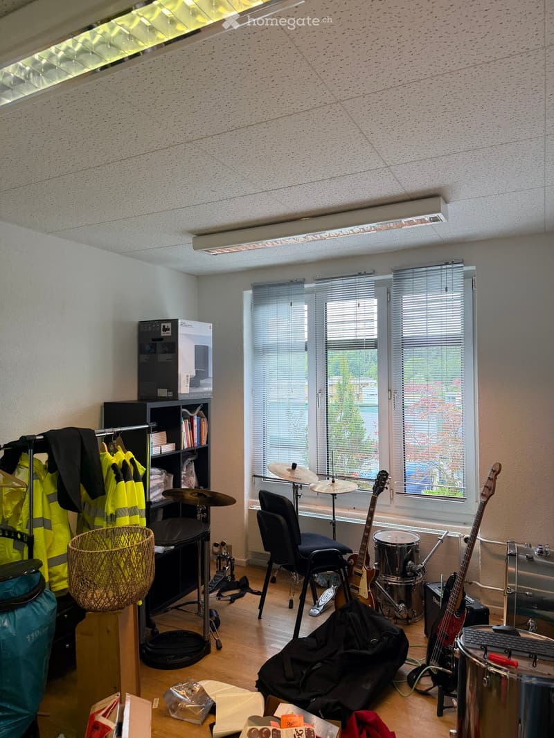 Furnished Room. A room with a drum set, guitars, a chair, a bookshelf, and a clothing rack. The room has large windows with blinds and a wooden floor.