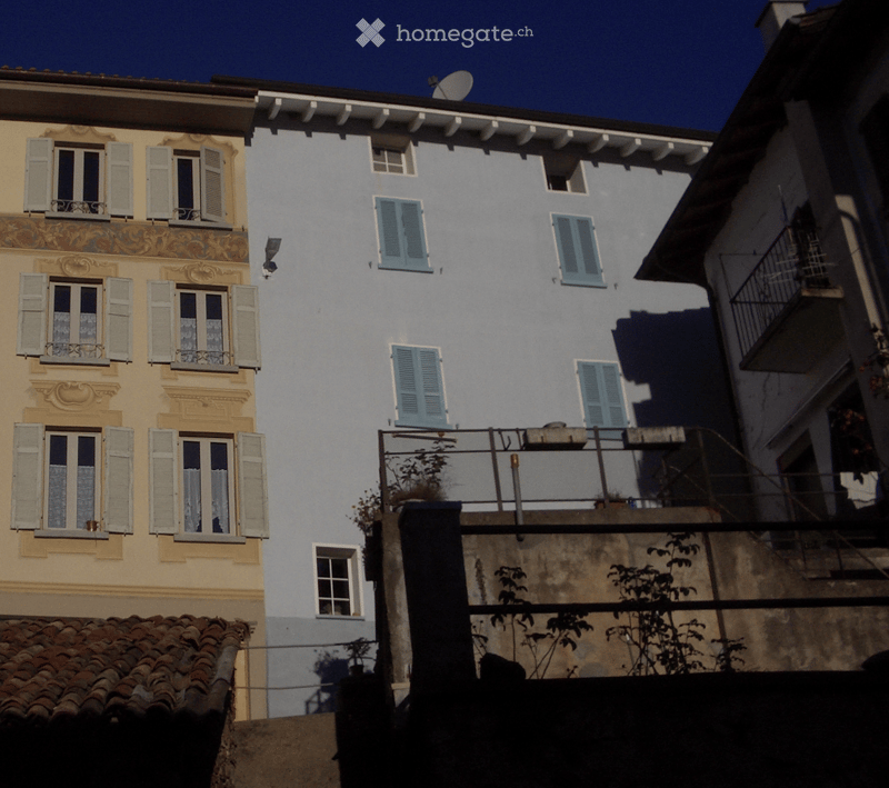 Building. Two-story residential building with light blue and cream-colored facades, multiple windows with shutters, and a small balcony on the upper floor.