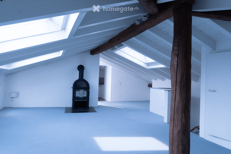 Unfurnished Room. An empty, bright room with white walls and a high, slanted ceiling. Features include exposed wooden beams, skylights, a black wood-burning stove, and a visible doorway leading to another space.