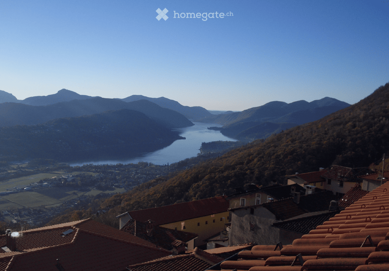 Building. Panoramic view of a town with red-tiled roofs, surrounded by lush green hills and a serene lake in the background.