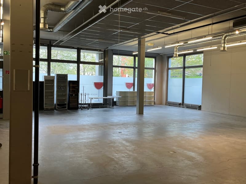 Unfurnished Room. Large, open, unfurnished room with concrete floors, exposed metal beams, and large windows allowing natural light. Features industrial-style lighting and a few small storage units and a table in the background.