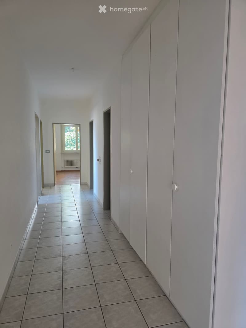 Empty hallway with white tiles, white walls, white cabinets, and a window with a view of trees.