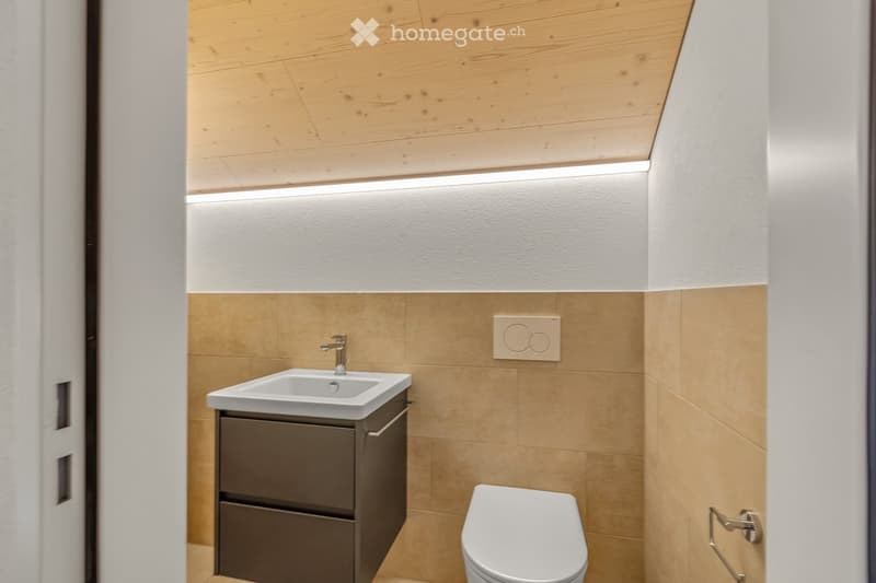 Bathroom. Modern bathroom with a white sink, dark grey cabinet, and a toilet. The walls are tiled in beige, and the ceiling is wooden with integrated lighting.