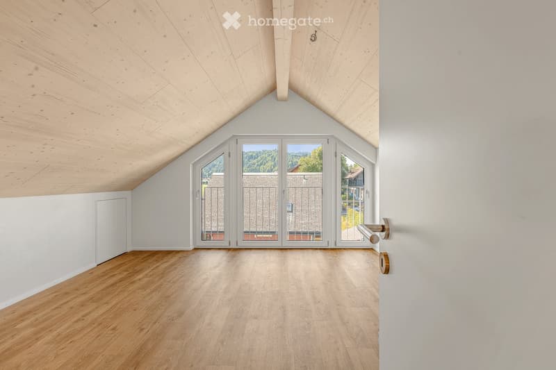 Unfurnished Room. An empty attic room with wooden flooring, white walls, and large windows offering a view of the outdoors. The ceiling has a sloped, wooden design.