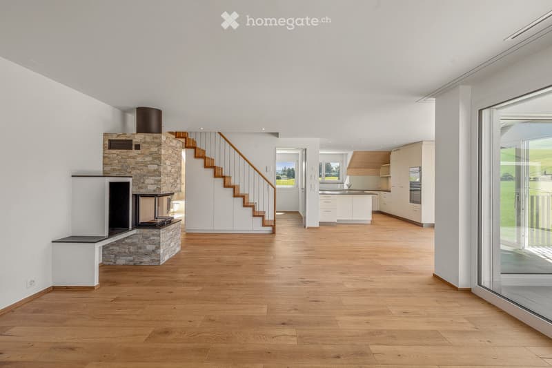 Furnished Room. Open-plan living space with wooden flooring, a stone fireplace, and a staircase leading to an upper level. The room features large windows and a view of the outdoors.