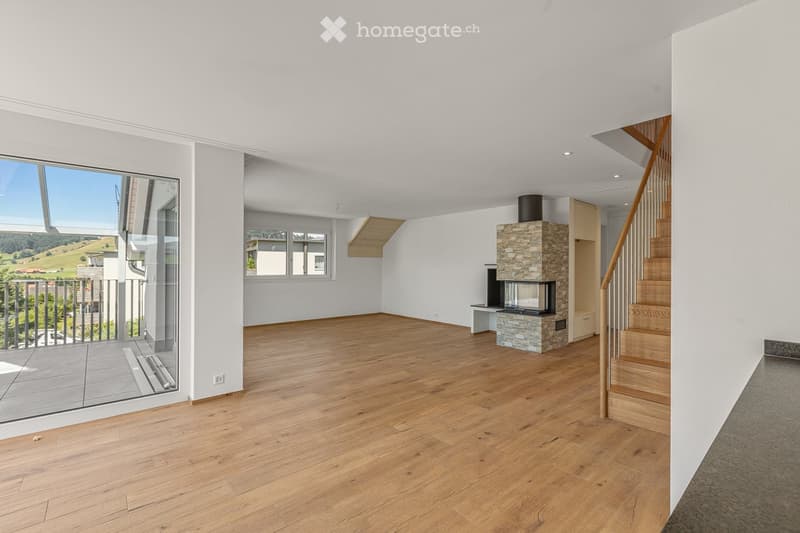 Furnished Room. Bright, open living space with wooden flooring, a stone fireplace, and a staircase leading to another level. Large windows offer a scenic view of the surrounding landscape.