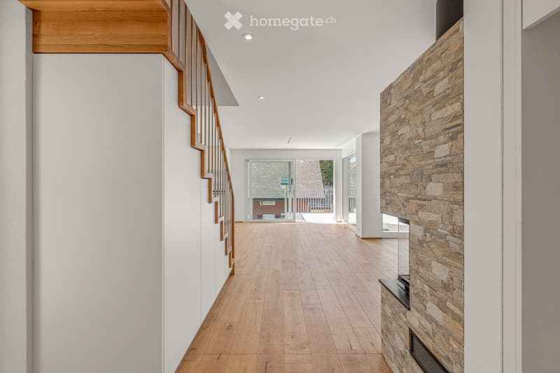 Corridor / Hallway. Modern hallway with wooden flooring, white walls, and a stone fireplace on the right side. A staircase with wooden railings is visible on the left, leading to an upper level. Large windows at the end provide natural light and a view of