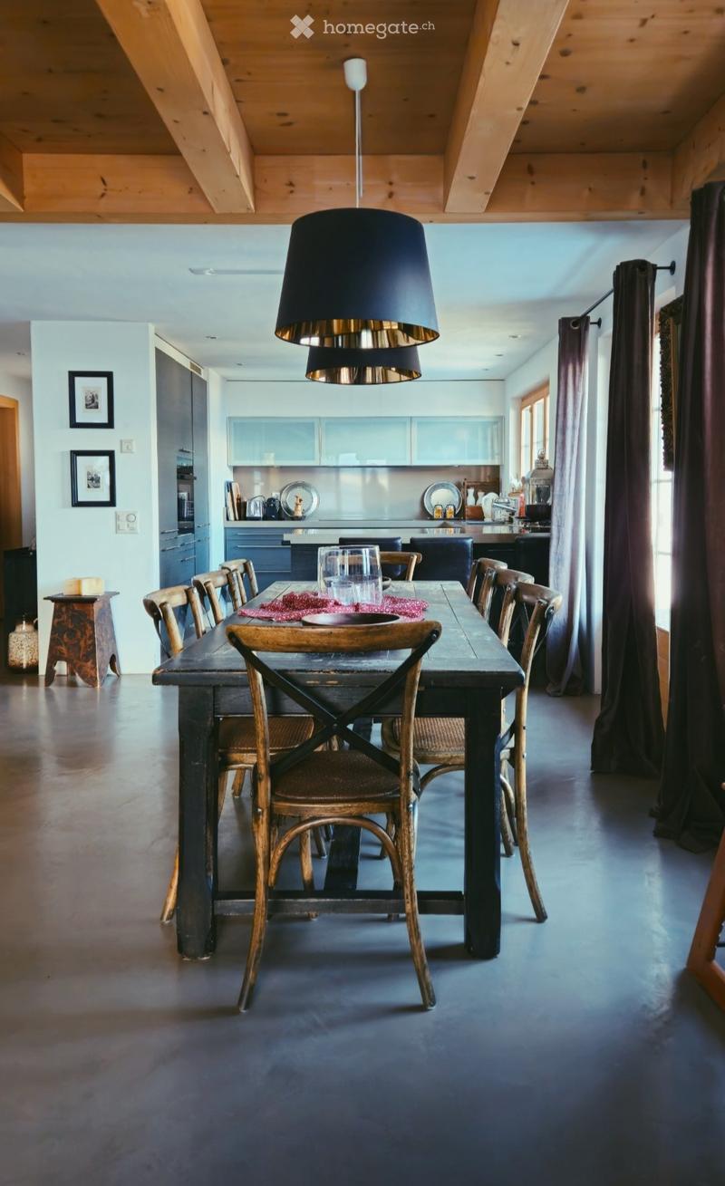 Dining room with wooden dining table and chairs, a large pendant light, wooden ceiling beams, kitchen, and wooden floor.