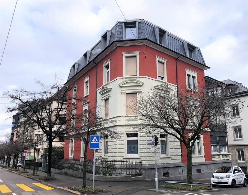 Building. A three-story residential building with red and white facade, multiple windows, and a grey roof with dormer windows. The building is situated on a street with trees, a pedestrian crossing sign, and a small car parked nearby.