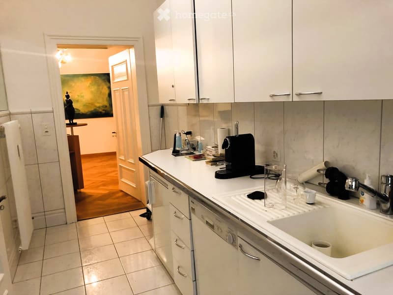 Kitchen. Modern kitchen with white cabinets, tiled floor, and a double sink. Coffee maker, cups, and other small items are on the counter. A doorway leads to another room with a painting visible.