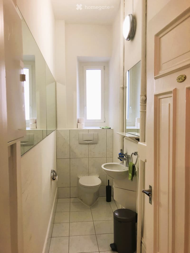 Bathroom. Compact bathroom with white tiled walls and floor, toilet, sink, mirror, and a small window above the toilet.