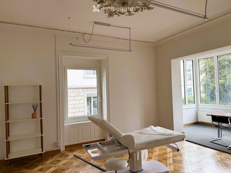 Furnished Room. Spacious room with wooden flooring, large windows, a treatment table, a stool, a shelf with decorative items, and a small seating area with a table and chairs.