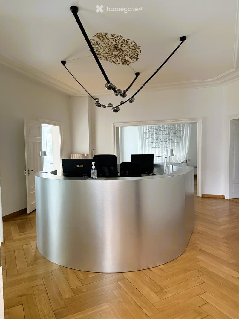 Furnished Room. Modern reception area with a sleek, curved white reception desk, a laptop, chairs, lamps, and a decorative chandelier.