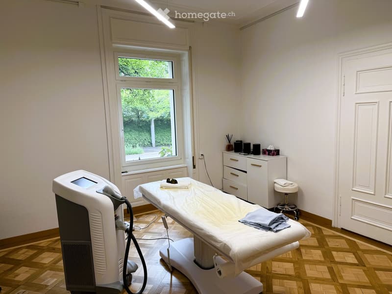 Furnished Room. A well-lit, clean treatment room with a wooden floor, large windows, a treatment table, a white cabinet, a small stool, and a medical device.
