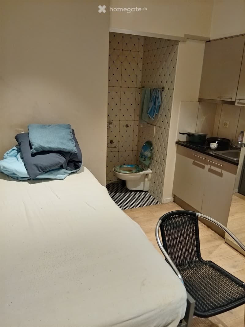 Single bed with pillows, chair, small bathroom with toilet, kitchen with sink and stove