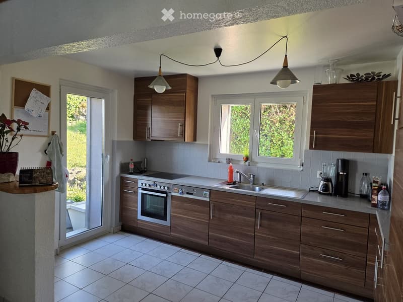 Kitchen. Modern kitchen with wooden cabinets, stainless steel appliances, and a large window overlooking greenery. Includes an oven, sink, blender, coffee maker, and various bottles on the counter.