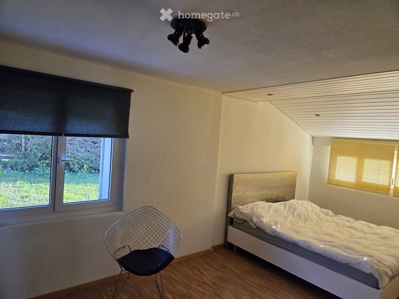 Bedroom. Bedroom with a single bed, a modern wireframe chair, and large windows with blinds. The room has light-colored walls and wooden flooring.