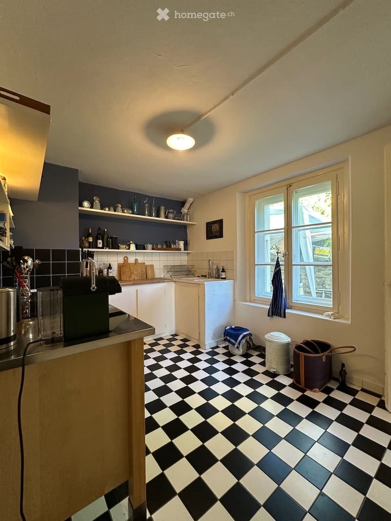 Kitchen. The kitchen features a black and white checkered floor, white cabinets, and a dark blue accent wall. Visible items include a sink, faucet, shelves with bottles and cups, a cutting board, a coffee machine, and a trash bin near the window.