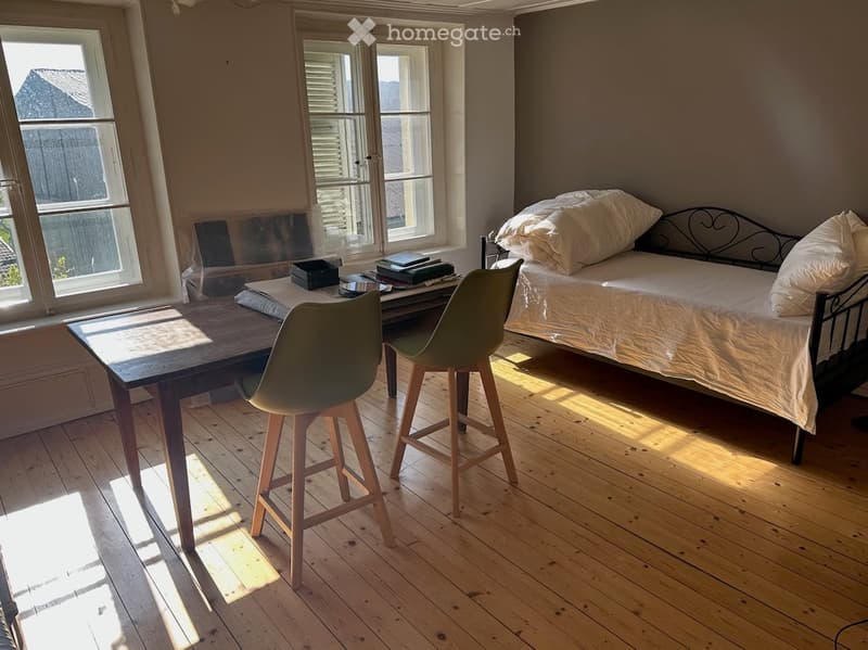 Bedroom. Bright bedroom with wooden flooring, large windows, a bed with white bedding, a wooden table with a chair and stool, and a small shelf with books and a box.