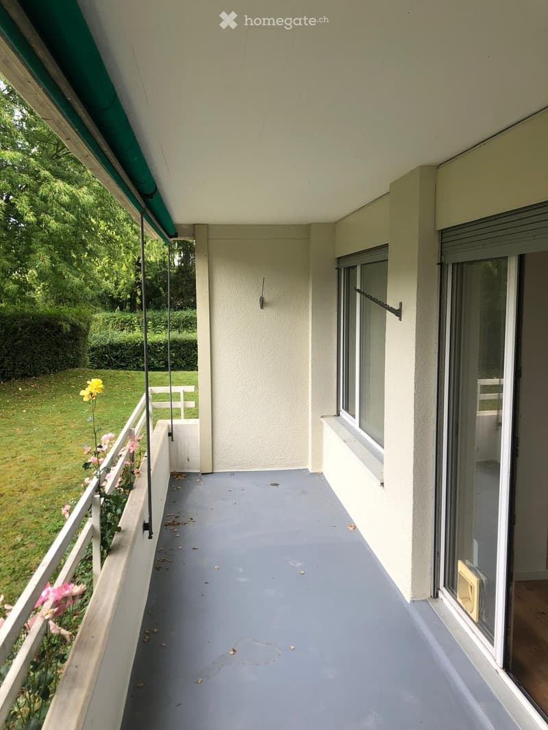 Balcony / Terrace. Balcony with grey flooring, white railing, and a view of a green lawn and trees. There are pink and yellow flowers along the railing.