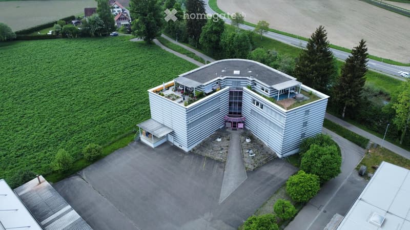 Building. Modern, multi-story building with a distinctive curved roof and white facade, surrounded by greenery and a paved parking area.