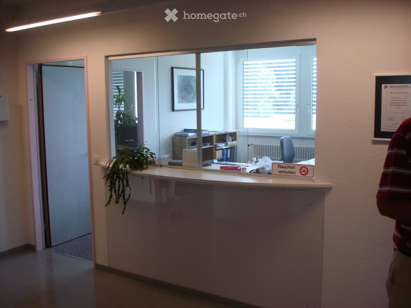 Furnished Room. Reception area with a desk, chair, plant, framed picture, and office supplies visible through a glass partition.