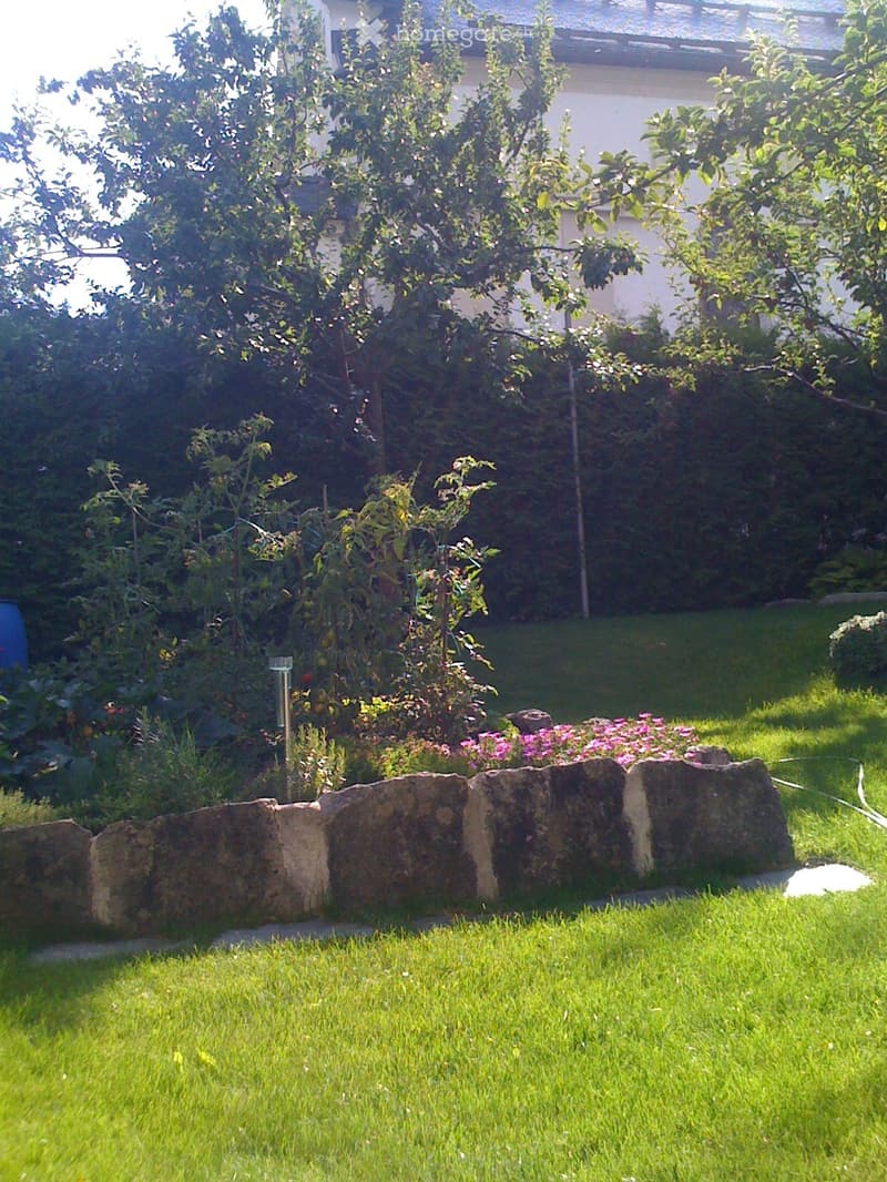 lawn, shrubs, trees, garden bed, hedge, stone wall, flowerbed, and a blue barrel in the background